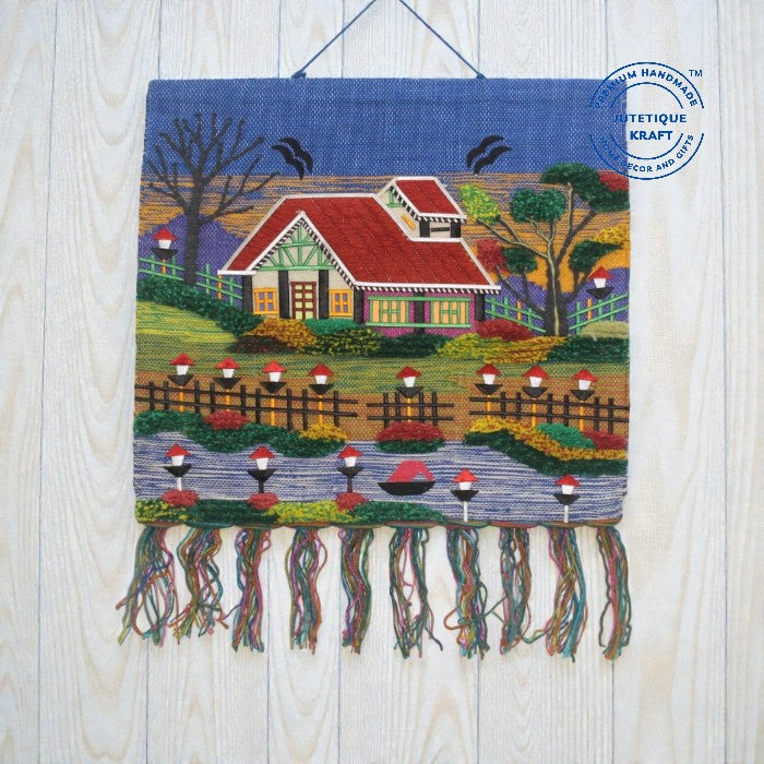Hand-embroidered cottage tapestry wall hanging named The Red Cabin Scenery, featuring a red-roofed house surrounded by green trees, birds, and decorative border fringes by Jutetique Kraft.